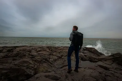 Man standing on rocky shore in Duluth, Minnesota, facing the lake under overcast sky.