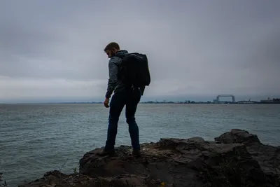 A man with a backpack stands on rocky shore, gazing over Duluth's waterfront.