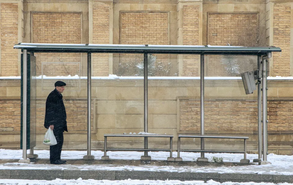 A man waits at a bus stop during a snowy winter day, holding a bag.