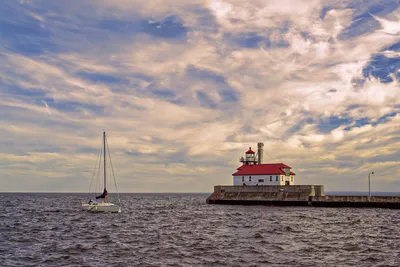 Duluth lighthouse with sailboat on a serene day under a dramatic sky.